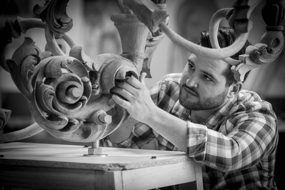Un hombre se concentra intensamente mientras talla o ensambla una escultura de madera ornamentada e intrincada, trabajando con las manos en una mesa. La imagen es en blanco y negro.