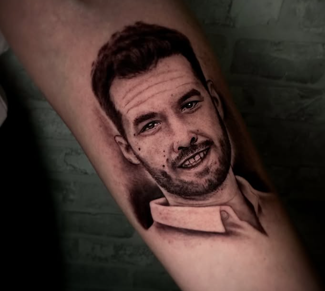 Tatuaje realista en negro y gris de un hombre sonriente con pelo corto y barba recortada en el antebrazo de una persona, con un fondo de sombras suaves y una camisa de cuello visible.