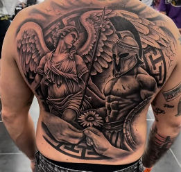 Un detallado tatuaje negro y gris cubre la espalda de una persona y representa a un ángel alado que sostiene un bastón y a un guerrero griego con casco, rodeado de intrincados dibujos y una mano que sostiene una flor en la parte inferior.