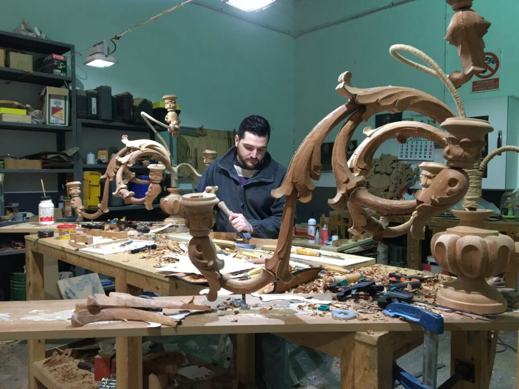 Un hombre trabaja en intrincadas tallas de madera en una desordenada mesa de taller, rodeado de herramientas, virutas de madera y grandes piezas decorativas de madera bajo brillantes luces cenitales.