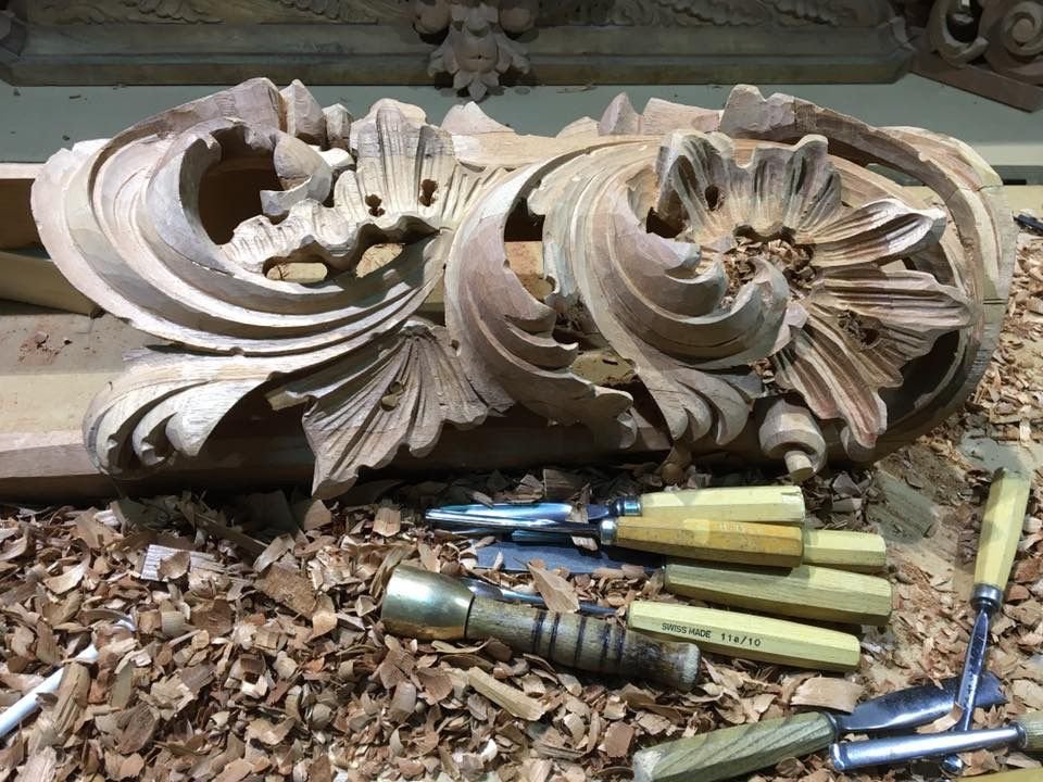 Una detallada talla de madera con intrincados diseños florales y volutas descansa sobre un banco de trabajo, rodeada de virutas de madera y varios cinceles y herramientas de talla.