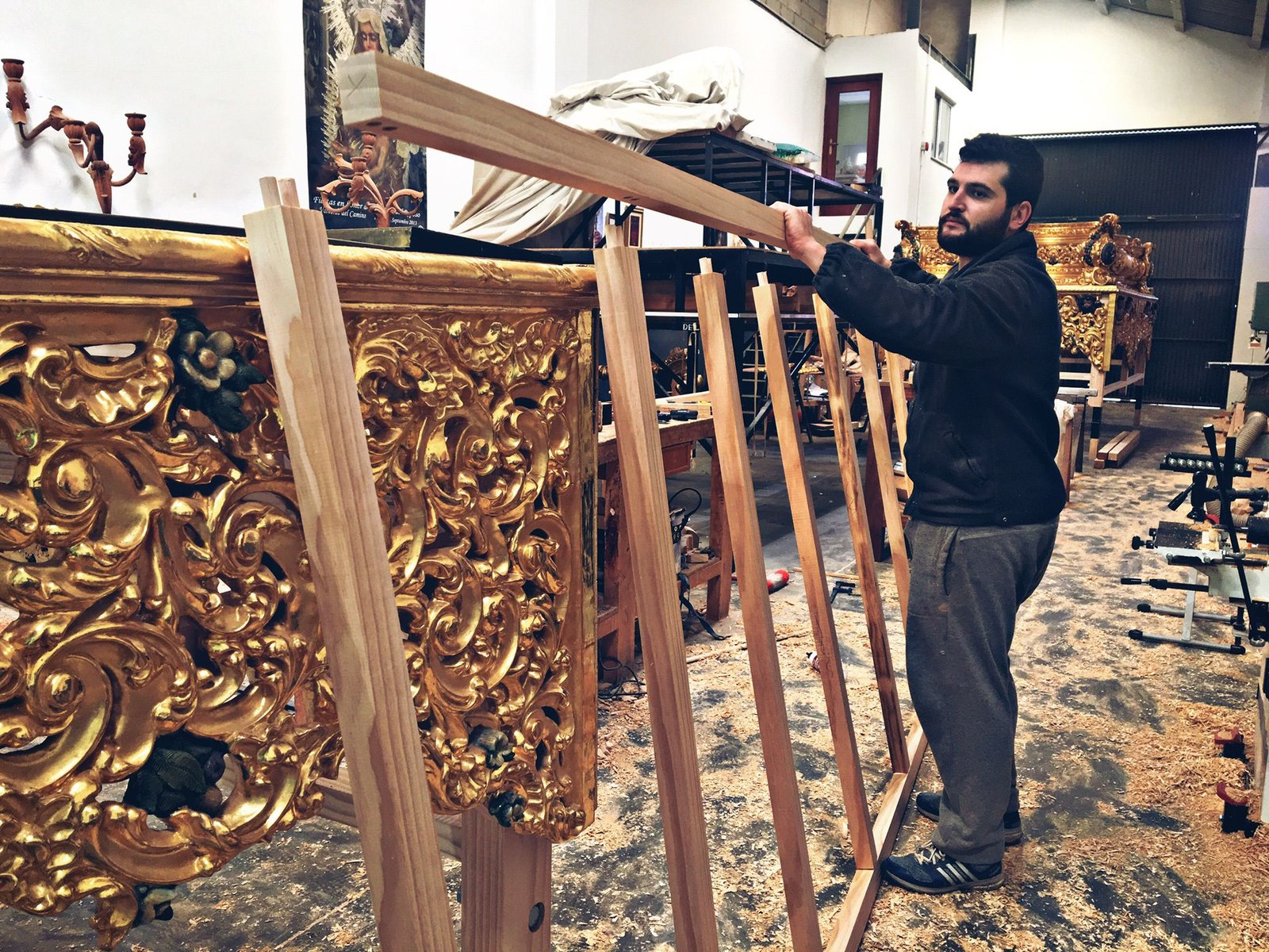 Un hombre se encuentra en un taller, trabajando con vigas de madera junto a una estructura dorada y ornamentalmente tallada. El suelo está cubierto de serrín y al fondo se ven herramientas y materiales diversos.