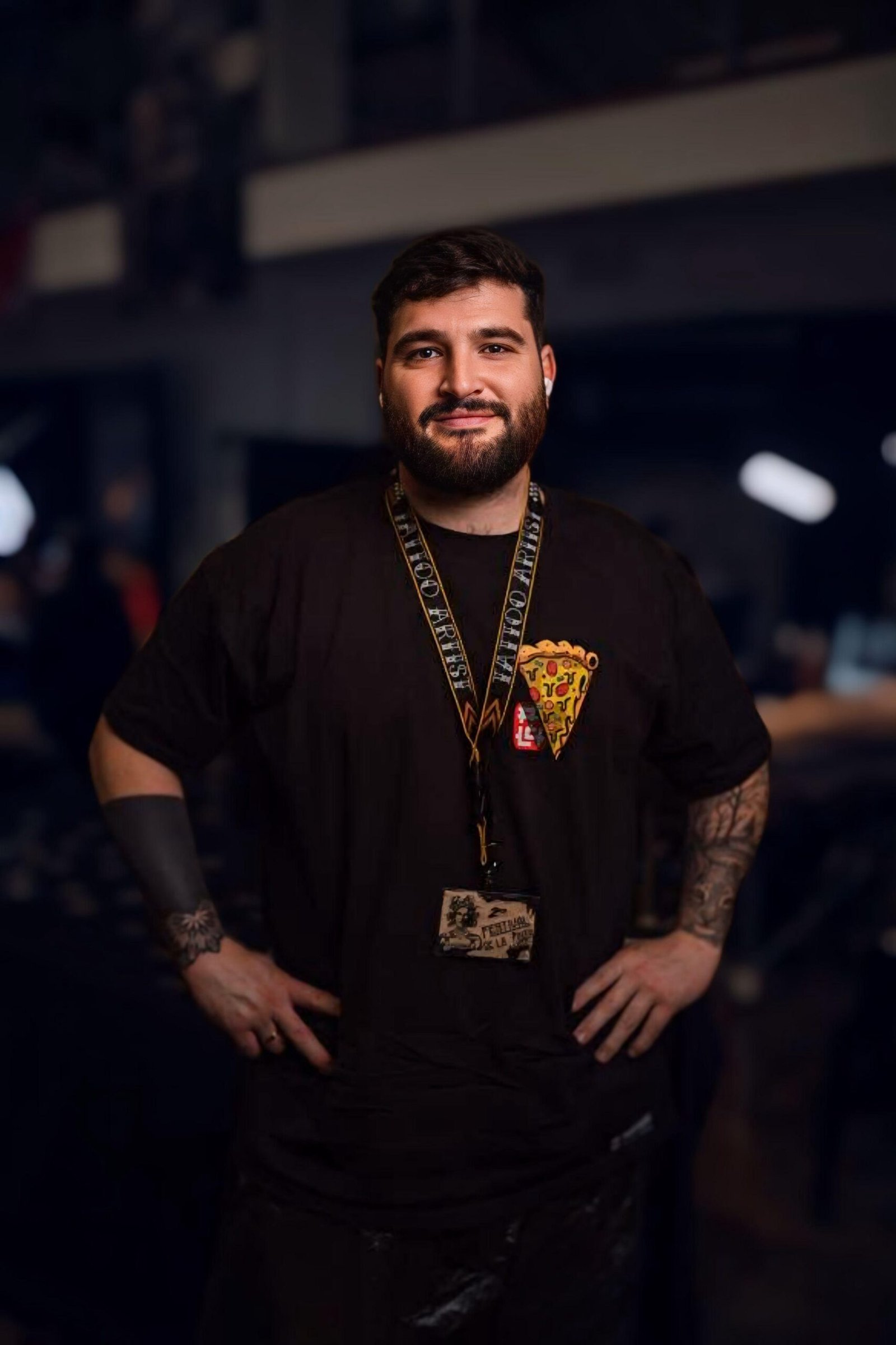 Un hombre con barba y tatuajes está de pie en el interior, sonriendo con las manos en las caderas. Lleva una camiseta negra con un gráfico de pizza y un cordón con una tarjeta de identificación alrededor del cuello. El fondo está poco iluminado y borroso.